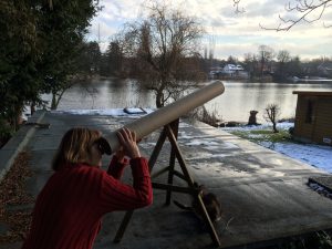 Die Blindgängerin auf dem Dach eines großen Gartenschuppens, neben einer verschneiten Wiese vor einem kleinen See. Sie steht vor einem dreibeinigen Stativ aus Holz. Darauf liegt eine sehr lange dunkle Pappröhre. Die Blindgängerin im roten Pullover schaut durch die Röhre wie durch ein Fernrohr in den Himmel.