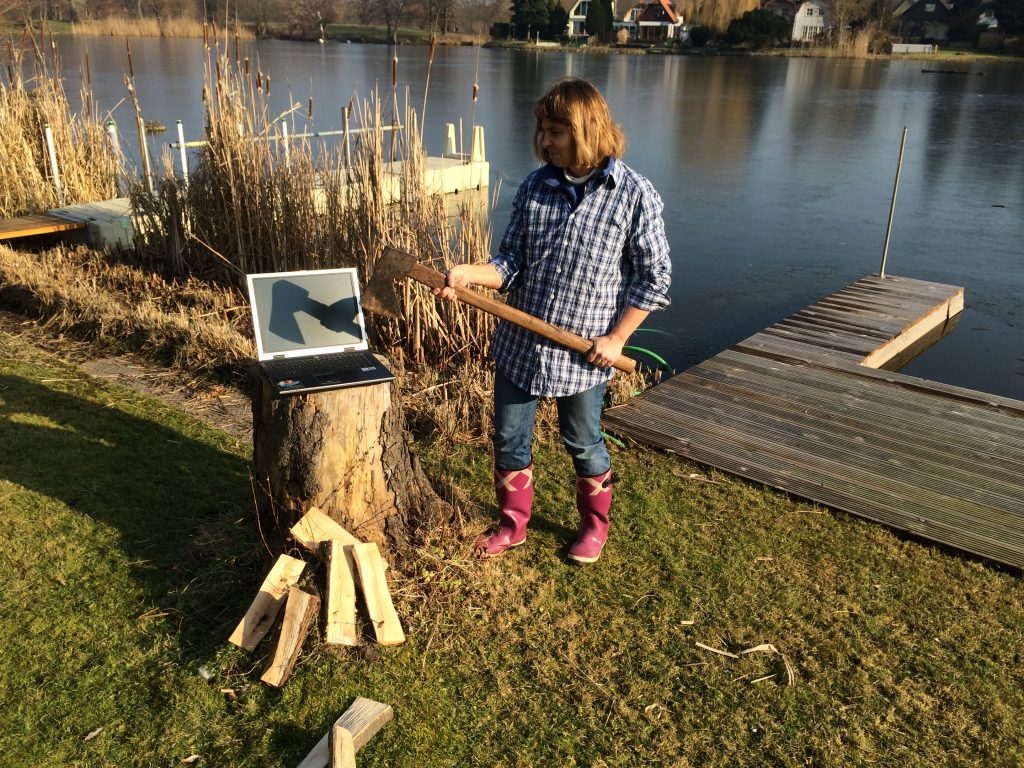 Auf einer Wiese am Seeufer, im Hintergrund kleine Bootsstege und Schilfgras. Auf einem Baumstumpf steht ein aufgeklappter Laptop, davor liegen Holzscheite. Die Blindgängerin im karierten Holzfällerhemd, Jeans und Gummistiefeln steht daneben und hält eine Axt. Auf dem Display des Laptops spiegelt sich der Schatten der Axt.