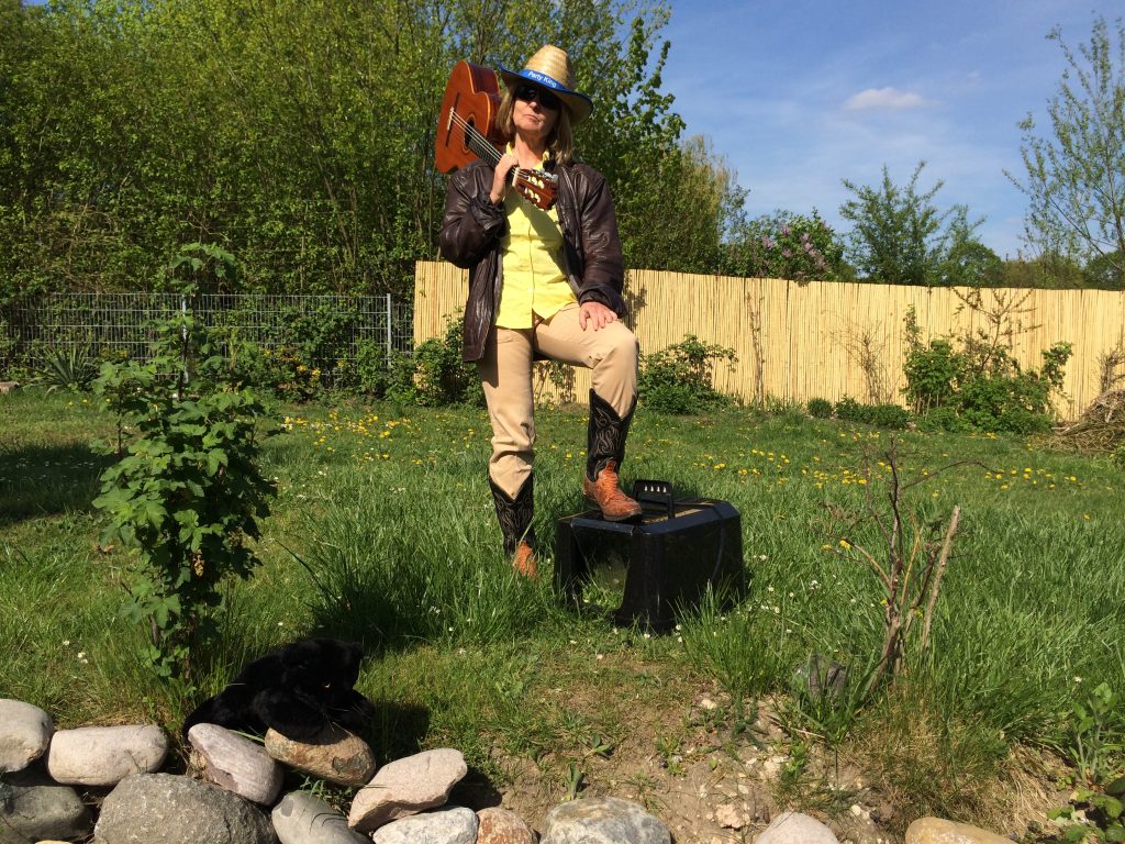 In einem verwilderten Garten steht die Blindgängerin im Sonnenlicht. Sie trägt einen hellen Cowboyhut, Lederjacke, beigefarbene Jeans und Cowboystiefel. Auf der Schulter hält sie eine Gitarre. Einen Fuß stellt sie auf den Deckel eines Katzenklos. Im Vordergrund ein paar helle Steine, auf denen eine schwarze Stoffkatze sitzt.