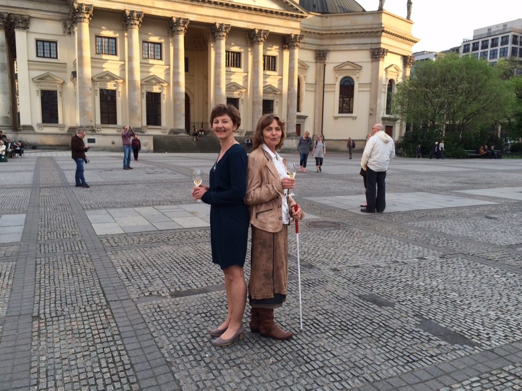 Auf dem Berliner Gendarmenmarkt vor dem Deutschen Dom. Zwei Frauen stehen Rücken an Rücken und lachen in die Kamera. Links Verena im dunklen Etuikleid, rechts die Blindgängerin im braunen Lederkostüm. Beide halten Sektgläser.