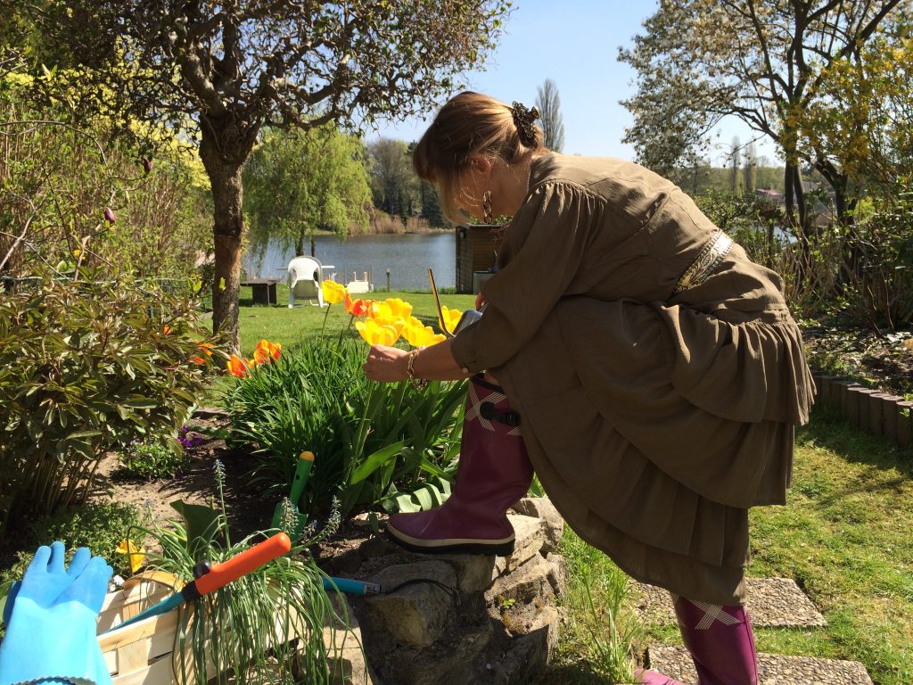 Die Blindgängerin beugt sich über ein Blumenbeet mit gelben Tulpen, in der rechten Hand ein kleines Gieskännchen. Sie hat die Haare zu einem Dutt gebunden und trägt lange goldene Ohrringe. Ihr wallendes Kleid ist kupferfarben und mit einem goldenen Gürtel gebunden. Sie trägt Gummistiefel. Neben ihr steht ein Korb mit Gartenwerkzeugen.