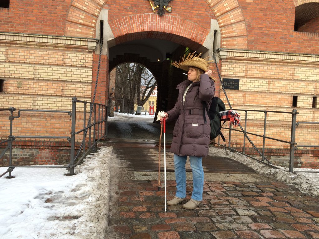 Vor einem offenen Burgtor mit Zugbrücke steht die Blindgängerin. Sie trägt einen ausgefransten Strohhut, eine lange dicke Winterjacke, Jeans und Stiefel. Auf ihren Rucksack ist eine zusammengerollte Wolldecke geschnallt. Die Blindgängerin hat eine Zigarette im Mund und hält neben ihrem weißen Langstock einen Wanderstock und eine weiße Muschel in der Hand.