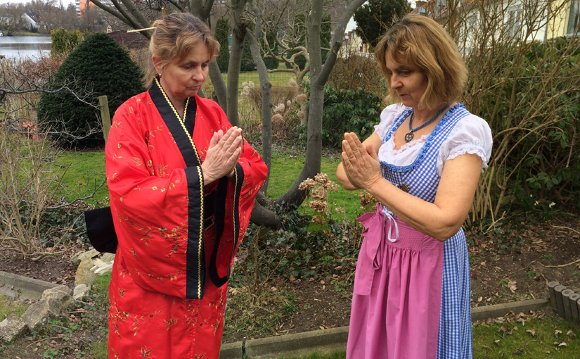 In einem Garten stehen sich zwei Frauen gegenüber, bei beiden handelt es sich um die Blindgängerin. Die linke hat das Haar mit einem Holzstäbchen hochgesteckt und trägt einen roten Kimono mit goldenen Stickereien und schwarzen Borten. Die rechte hat das schulterlange Haar offen und trägt über einer weißen Rüschenbluse ein blauweißkariertes Dirndl mit rosa Schürze. Beide neigen leicht die Köpfe und halten zur Begrüßung die Hände zusammengelegt vor die Brust.