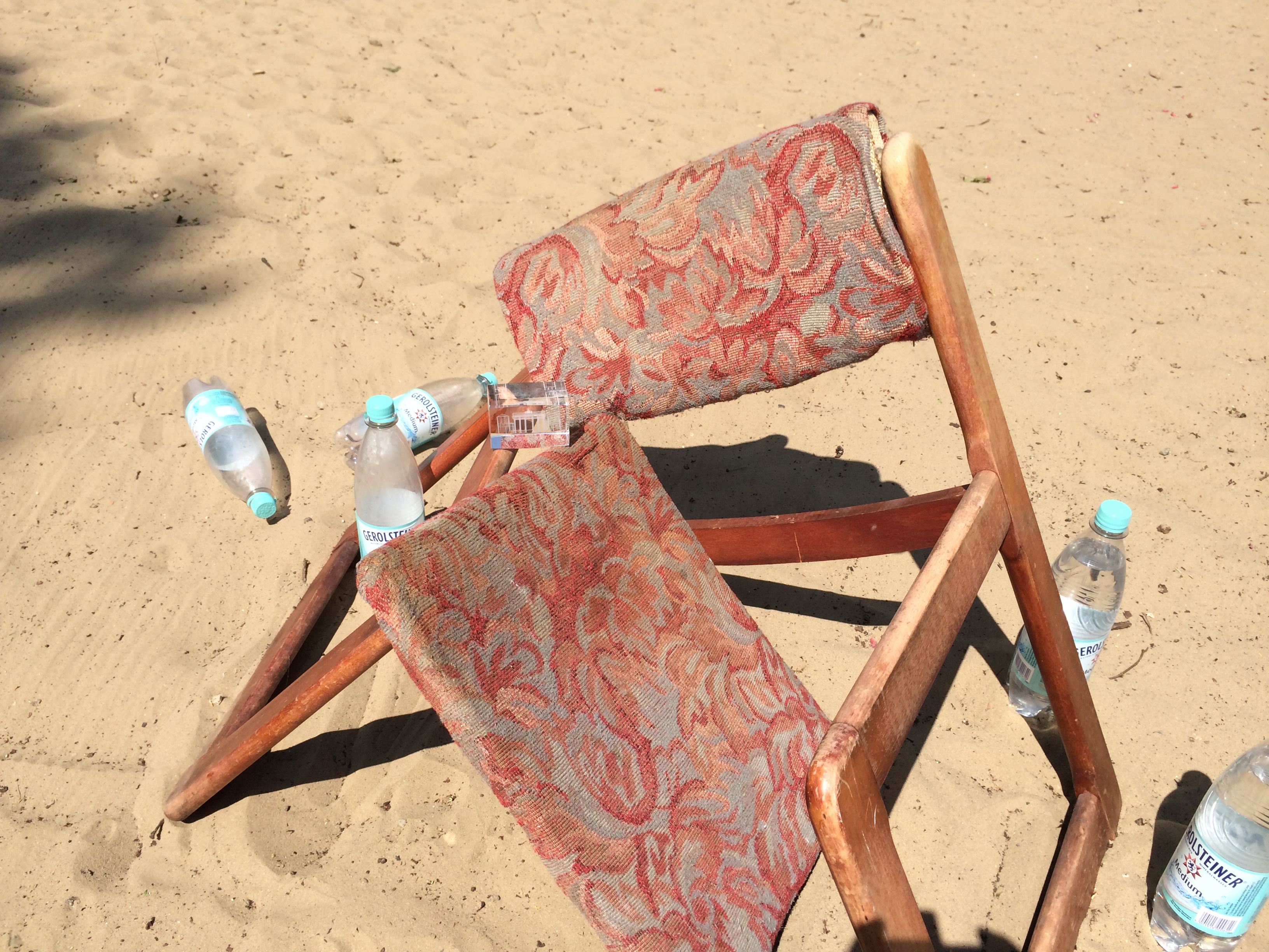 Auf einer Sandfläche steht ein brauner Holzstuhl. Rückenlehne und Sitzfläche sind mit Stoff bespannt, der in Rosatönen gemustert ist. Der Stuhl ist zerbrochen, die Beine nach außen geknickt, die Sitzfläche liegt mit einer Seite im Sand. Zu beiden Seiten des Stuhles stehen und liegen Wasserflaschen am Boden.