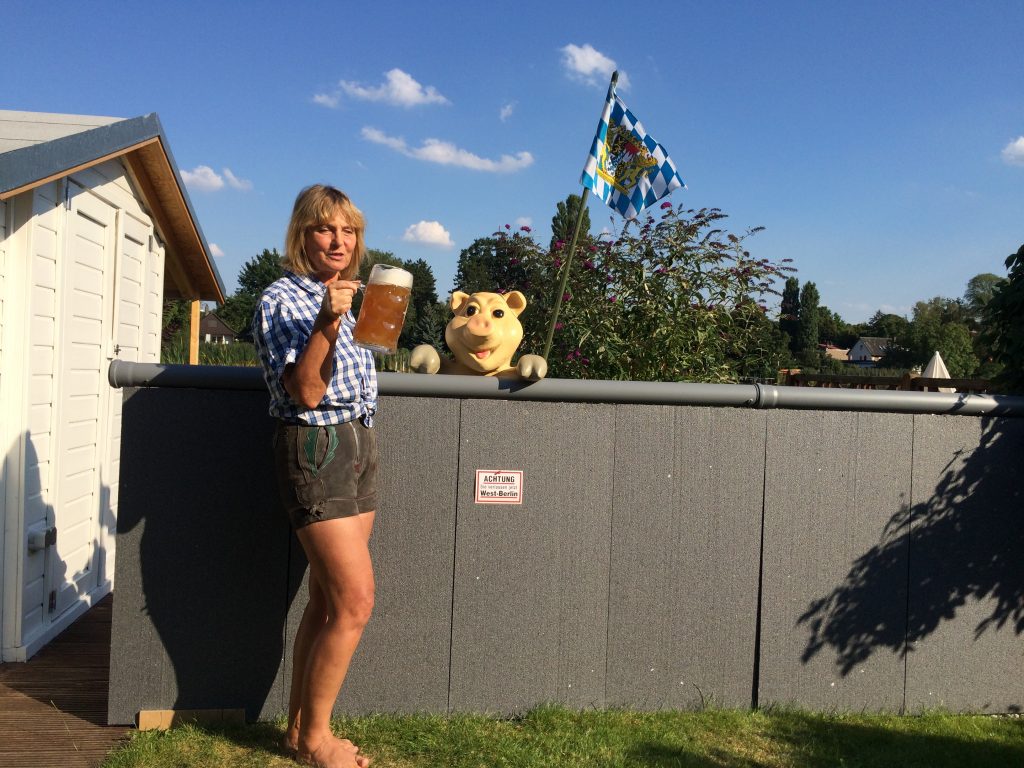 Auf einer Wiese unter blauem Himmel steht eine etwa einen Meter hohe Wand aus grauen Platten, obenauf liegt eine graue Röhre. An der Wand ein Schild: Achtung, Sie verlassen jetzt West-Berlin. Hinter der Mauer ist die bayerische Flagge gehißt und ein künstlicher Schweinskopf schaut über den Rand. Vor der Mauer steht die Blindgängerin. Sie trägt ein blauweißkariertes Shirt und eine kurze Trachtenlederhose. Mit einer vollen Maß Bier prostet sie in Richtung Kamera.