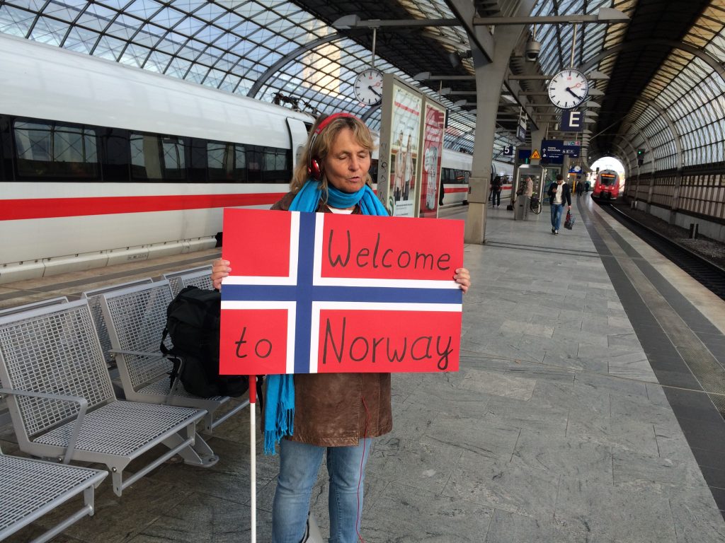 Auf dem Bahnsteig eines Fernbahnhofes, im Hintergrund ein haltender ICE. Die Blindgängerin hat rote Kopfhörer auf. Sie trägt einen blauen Schal, Lederjacke und Jeans. In den Händen hält sie ein großes Schild. Es zeigt die norwegische Nationalflagge mit der Aufschrift: Welcome to Norway.