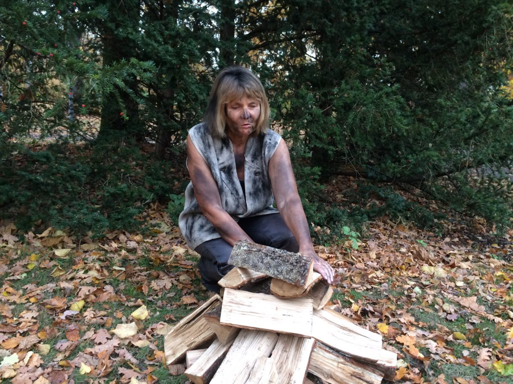 Vor einem Waldrand stapelt die rußverschmierte Blindgängerin Holzscheite zu einem Haufen.