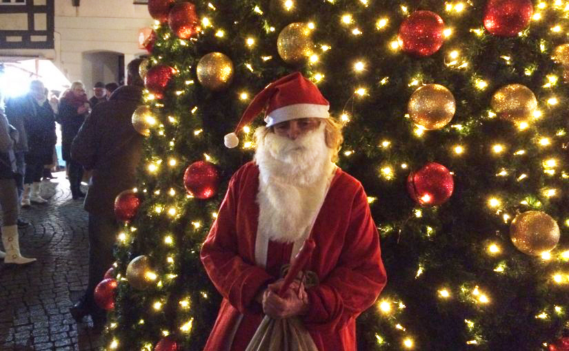 Die Blindgängerin als Weihnachtsmann verkleidet vor einem großen Weihnachtsbaum, an dem rote und goldene Kugeln glitzern.
