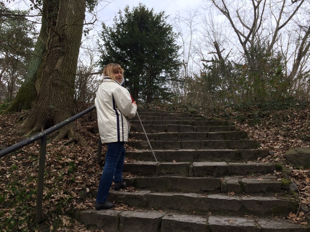 Die Blindgängerin in weißer Regenjacke beim Besteigen einer Steintreppe. Es liegt herbstliches Laub auf dem Waldboden. In der rechten hält sie ihren Langstock, in der linken das metallene Geländer. Über ihre Schulter dreht sie sich zur Kamera um.