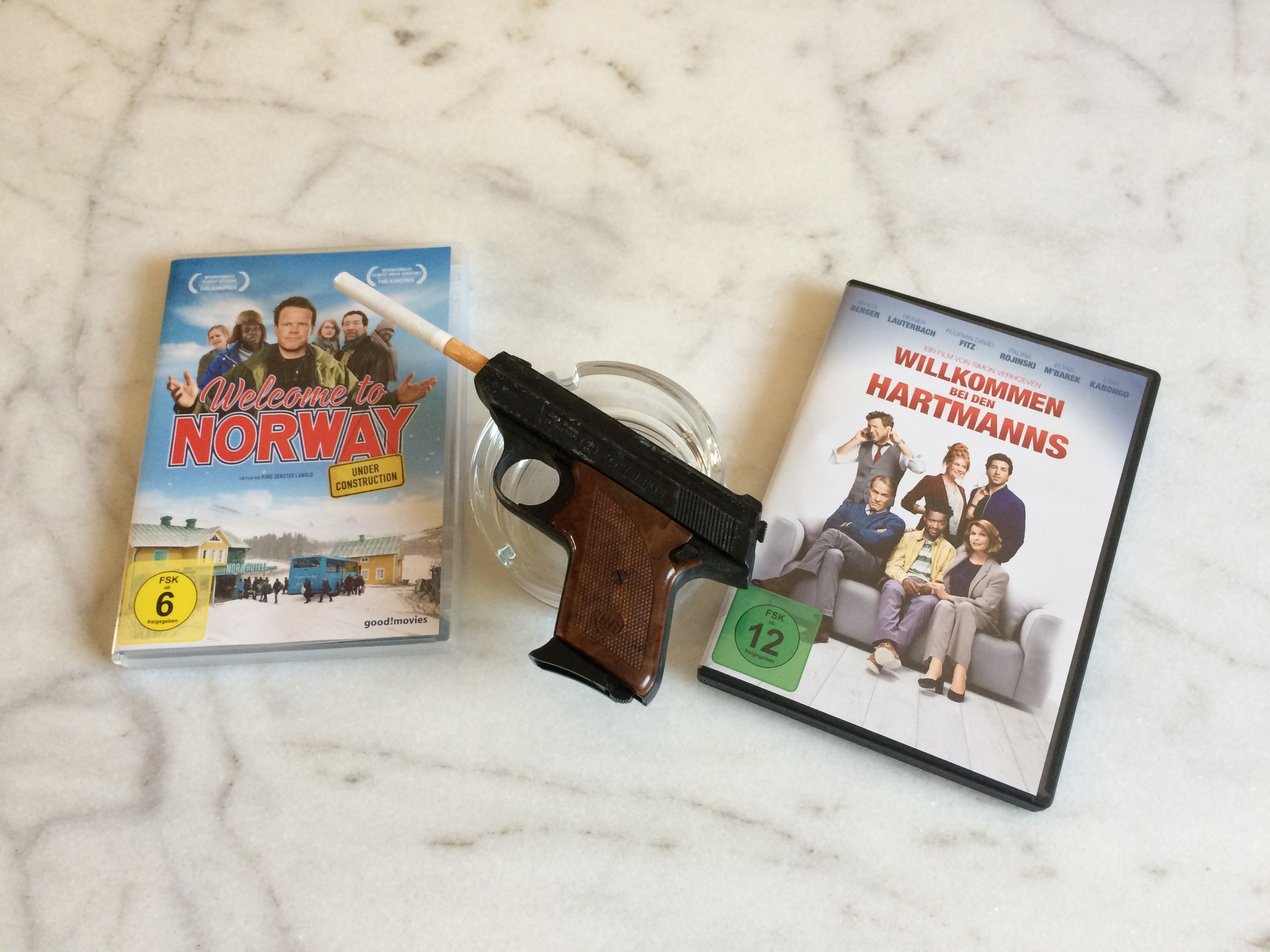 Zwei DVDs: Links des Films "Welcome to Norway", rechts von "Willkommen bei den Hartmanns". In der Mitte eine Pistole mit Filterzigarette im Lauf auf einem leeren Aschenbecher.