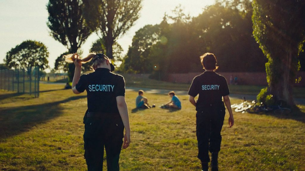 Filmstill aus "Tiger Girl": Die beiden Tigergirls in schwarzen Uniformen mit der Aufschrift "Security" auf dem Rücken laufen über eine Wiese im Park. Die vordere trägt ihr blondes langes Haar zu einem Zopf, darüber ein Basecap. Im Hintergrund sitzen zwei Leute im Gras.