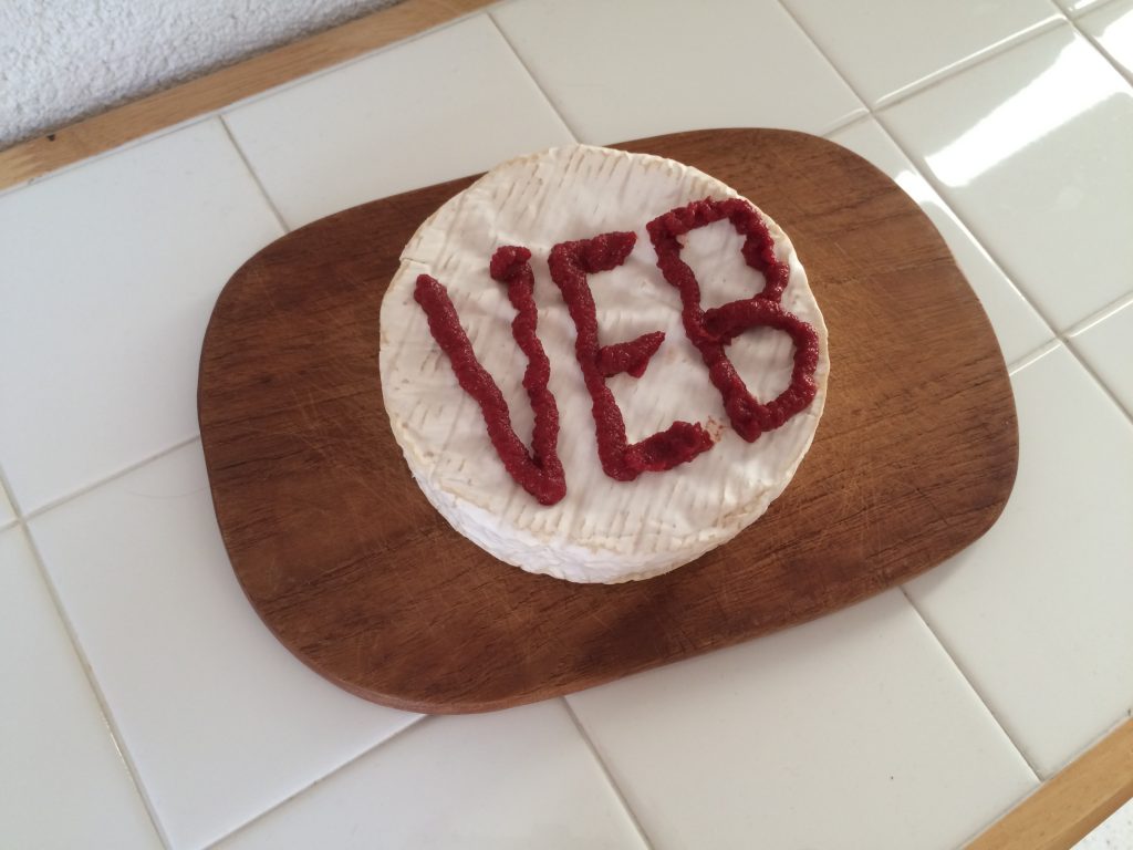 Auf einem Holzbrett ein kreisrunder Camembert, darauf steht mit Tomatenmark in großen Lettern V E B geschrieben