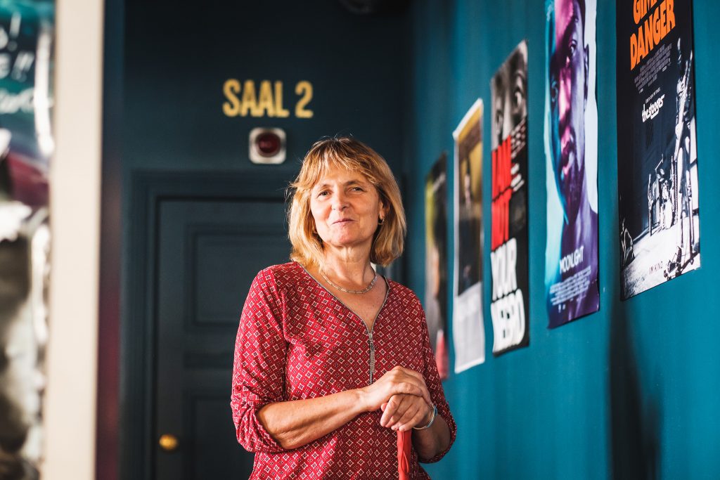 Barbara Fickert im Vorraum zu einem Kino. Hinter ihr steht in goldenen Lettern über einer Tür "Saal 2", zu ihrer Linken sind Filmplakate an einer blauen Wand angebracht. Barbara Fickert trägt ein rot gemustertes Oberteil und lächelt in die Kamera.