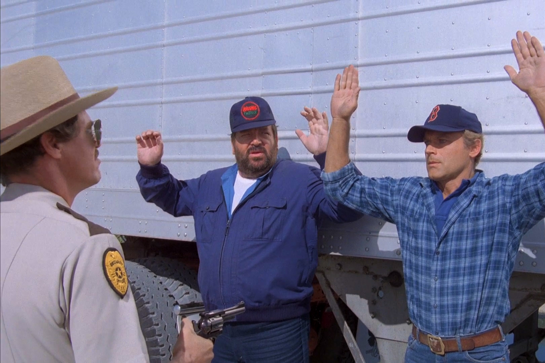 Bus Spencer und Terence Hill in einer Filmszene. Sie stehen mit erhobenen Händen vor einem LKW, während ein Polizist die Pistole auf sie richtet.