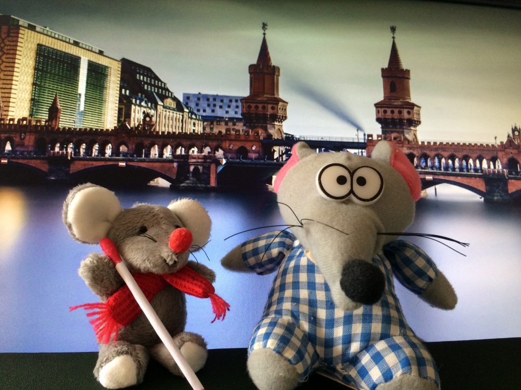 Zwei Mäuse als Stofffiguren vor der Berliner Oberbaumbrücke. Die kleinere Maus trägt einen roten Schal. Sie hält einen weißen Langstock. Die größere Maus schielt. Sie hat geknickte Ohren und eine sehr große gebogene Nase.