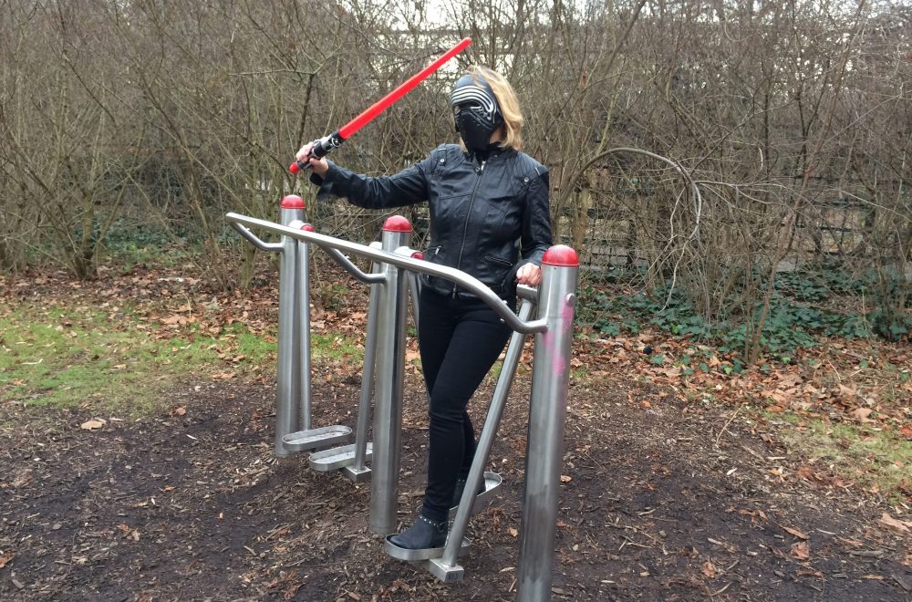 Die Blindgängerin steht auf einem Skywalker, einem Fitnessgerät, in einem Park. Sie trägt schwarze Kleidung und die schwarze Maske des Kylo Ren. Mit dem rechten Arm schwingt sie ein rotes Lichtschwert.