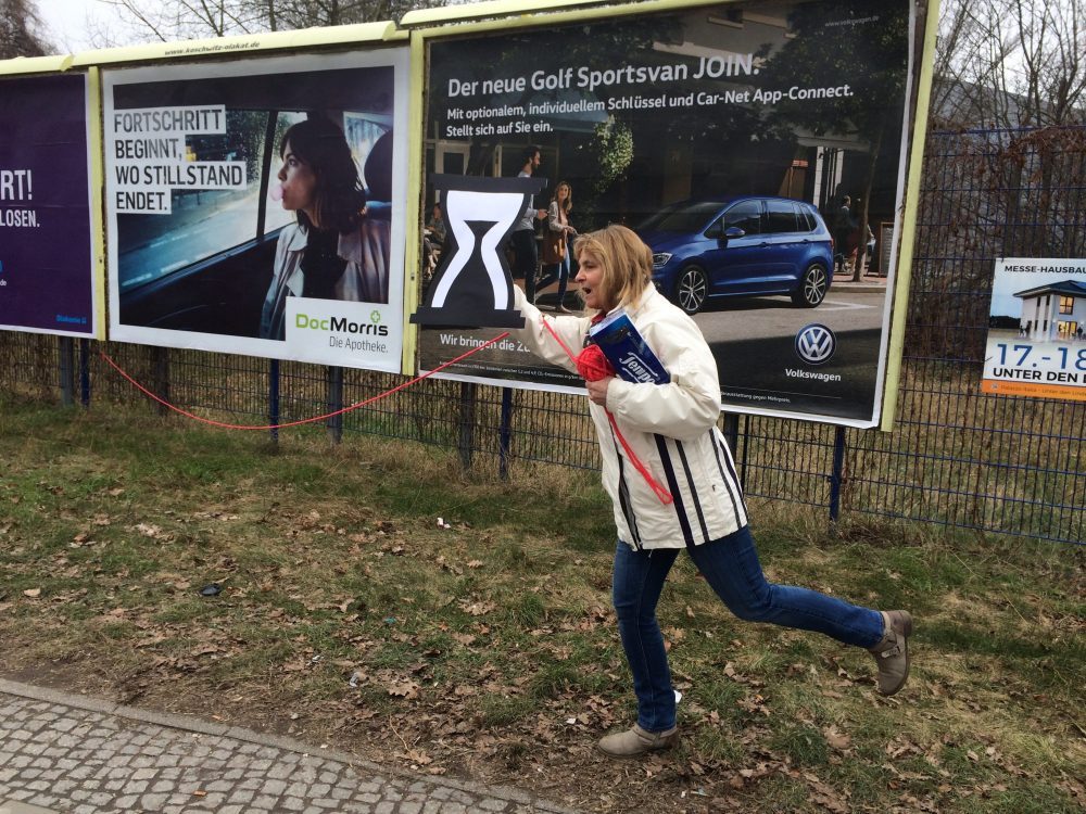 Die Blindgängerin im Laufschritt vor drei großen Werbetafeln. Von einer spannt sich ein roter Faden zu ihr, das Knäuel hält sie in der linken Hand. Unter dem Arm trägt sie ein großes Paket Papiertaschentücher. In der rechten Hand hält sie ein aus Pappe geschnittenes Stundenglas, eine Sanduhr.