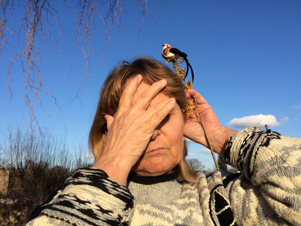 Die Blindgängerin in der Haltung der Skulptur "Die Lauschende". Vor einem blauen Himmel hält sie die rechte Hand vor Augen und Stirn, die Linke legt sie lauschend hinter das Ohr. In der linken Hand hält sie einen Zweig, auf dem ein kleiner bunter Vogel sitzt.