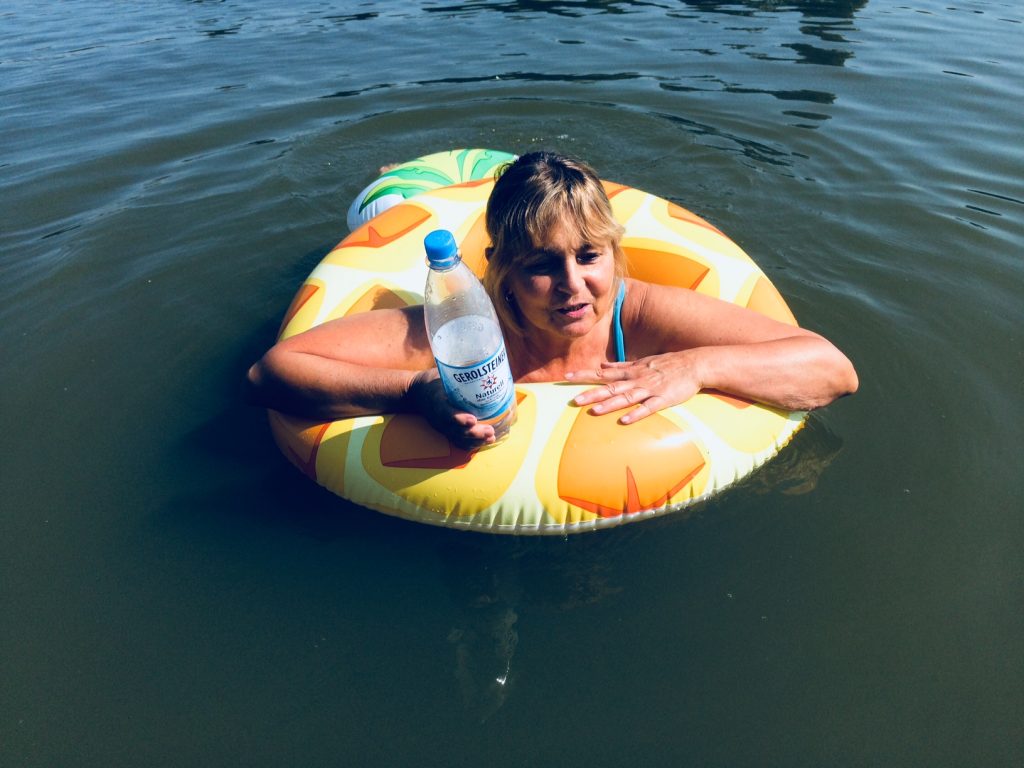 Die Blindgängerin schwimmt im Wasser mit einem aufblasbaren Schwimmring, der das Muster einer Ananas hat. In der rechten Hand hält sie eine Wasserflasche.