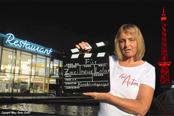 Die Blindgängerin hält eine Filmklappe in den Händen. Sie trägt ein weißes T-Shirt mit dem Aufdruck „And Action“. Im Hintergrund das Palais am Funkturm und der Funkturm hell erleuchtet vor dem schwarzen Nachthimmel.