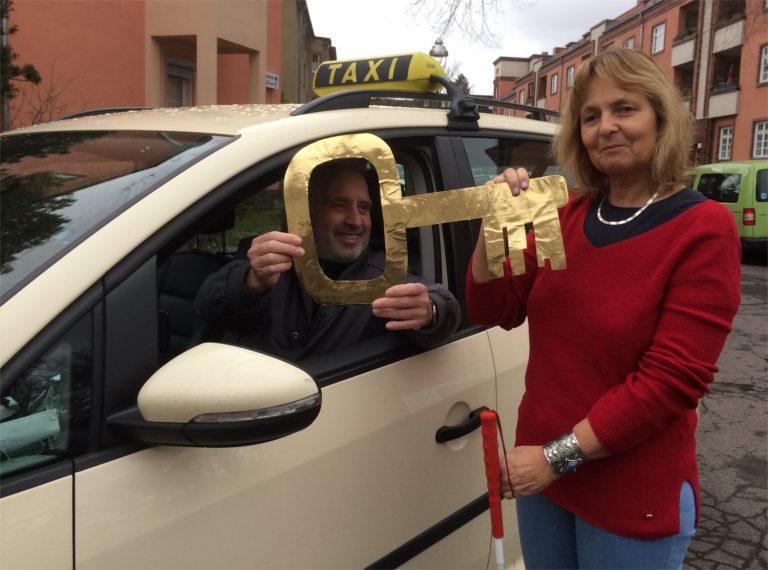 Die Blindgängerin im roten Pulli und Jeans steht neben einem Berliner Taxi. Der Taxifahrer hinterm Steuer reicht ihr lächelnd einen großen goldenen Schlüssel durchs Seitenfenster.