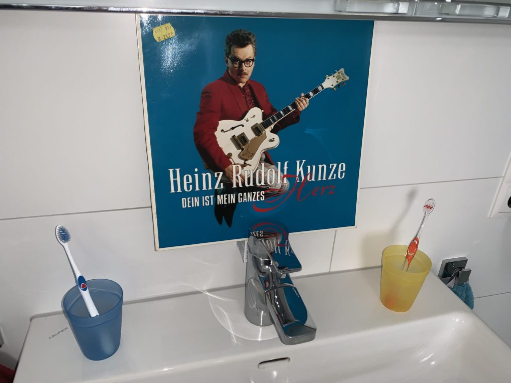 Auf einem Badezimmer-Waschbecken stehen links und rechts außen zwei Zahnputzbecher mit Zahnbürsten darin. In der Mitte über dem Wasserhahn hängt das Cover einer LP. Darauf vor dunkelblauem Hintergrund Heinz Rudolf Kunze. Er trägt ein rotes Jackett und springt in die Luft. Dabei hält er eine weiße halbakustische Gitarre. Der Titel der LP lautet "Dein ist mein ganzes Herz".