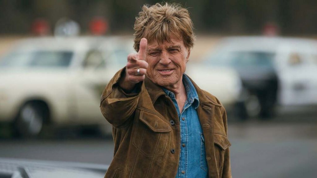 Robert Redford in blauem Jeanshemd und brauner Wildlederjacke. Die Haare zerzaust, der Ausdruck zwischen Lächeln und Grinsen. Mit der erhobenen rechten Hand formt er eine Pistole nach. Sie zielt direkt auf den Betrachter.