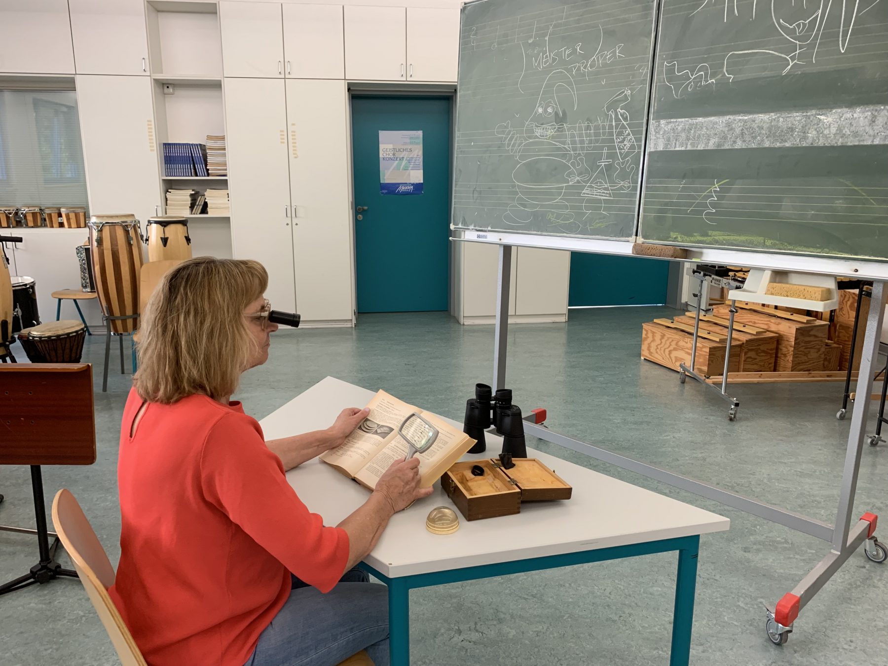 Die Blindgängerin sitzt in einem Schulraum an einem Tisch. Vor ihr eine Kreidetafel. Die Blindgängerin trägt eine Lupenbrille, vor ihr auf dem Tisch ein Schulbuch, ein Fernglas und zwei Leselupen.