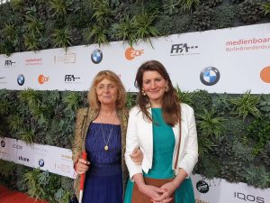 Die Blindgängerin und ihre Begleitung Lena stehen vor langen weißen Schildern mit den Logos der Sponsoren der Verleihung des Deutschen Filmpreises. Im Hintergrund eine grüne Hecke. Lena hat langes braunes Haar und trägt ein türkisfarbenes Kleid mit einer weißen Jacke. Die Blindgängerin trägt ihr mittelblondes Haar schulterlang. Einen dunkelblauen Rock und ein gleichfarbiges Top kombiniert sie mit einer kupfergoldenen Jacke mit aufgenähten blauen Glasperlen.