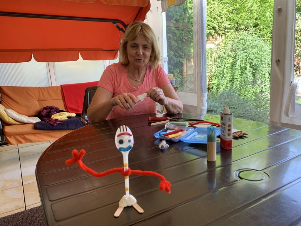 Die Blindgängerin sitzt mit einer Flasche Klebstoff in den Händen an einem Terrassentisch. Vor ihr liegt Bastelmaterial und daneben steht Forky, die Filmfigur, aus einem Göffel gebastelt.