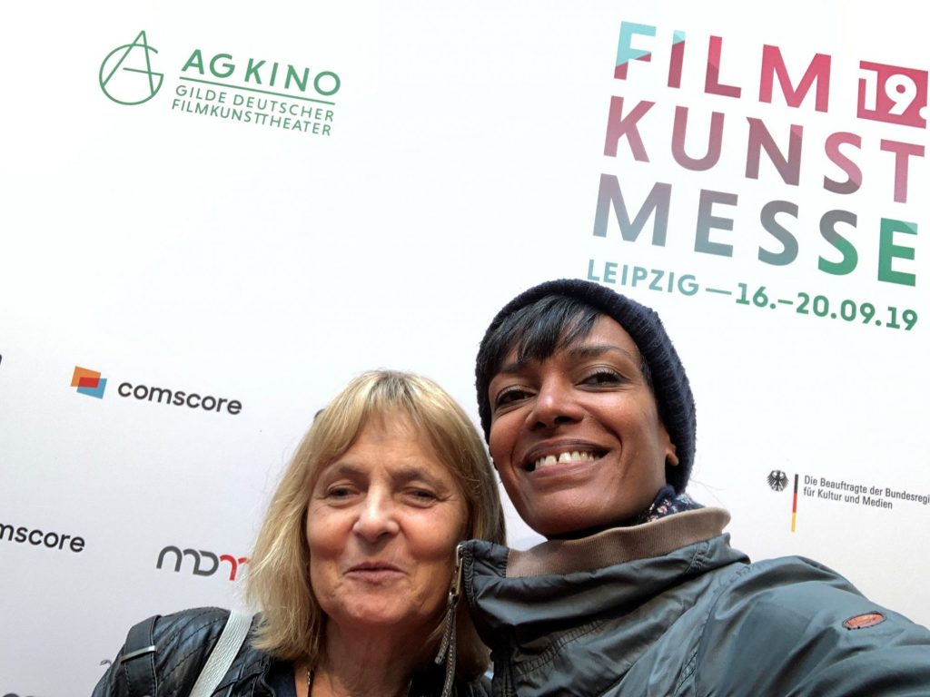 Ein Selfie zeigt Seneit Debese von der Greta und Starks App und die Blindgängerin. Sie stehen vor einem großen Plakat der 19. Filmkunstmesse Leipzig 2019.