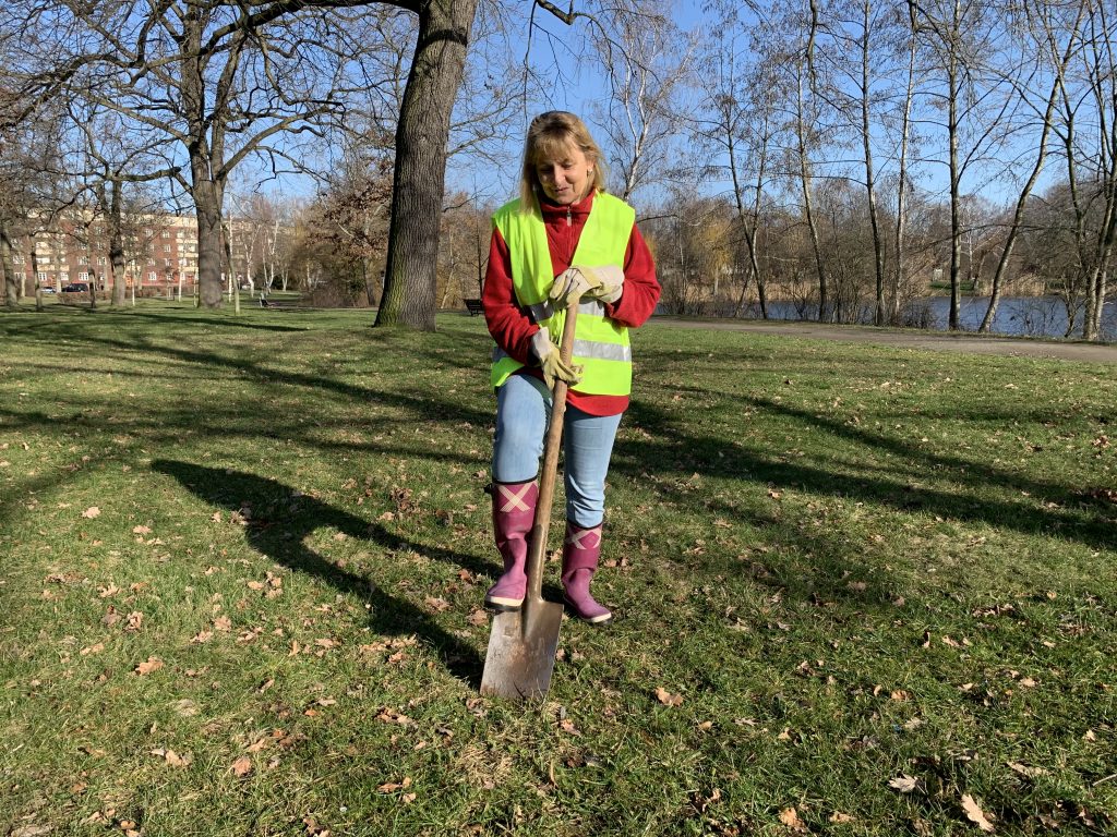 Die Blindgängerin steht auf einer sonnigen Wiese im Park. Sie trägt Gummistiefel, eine gelbe Warnweste und Arbeitshandschuhe. Sie sticht einen Spaten in den Boden.