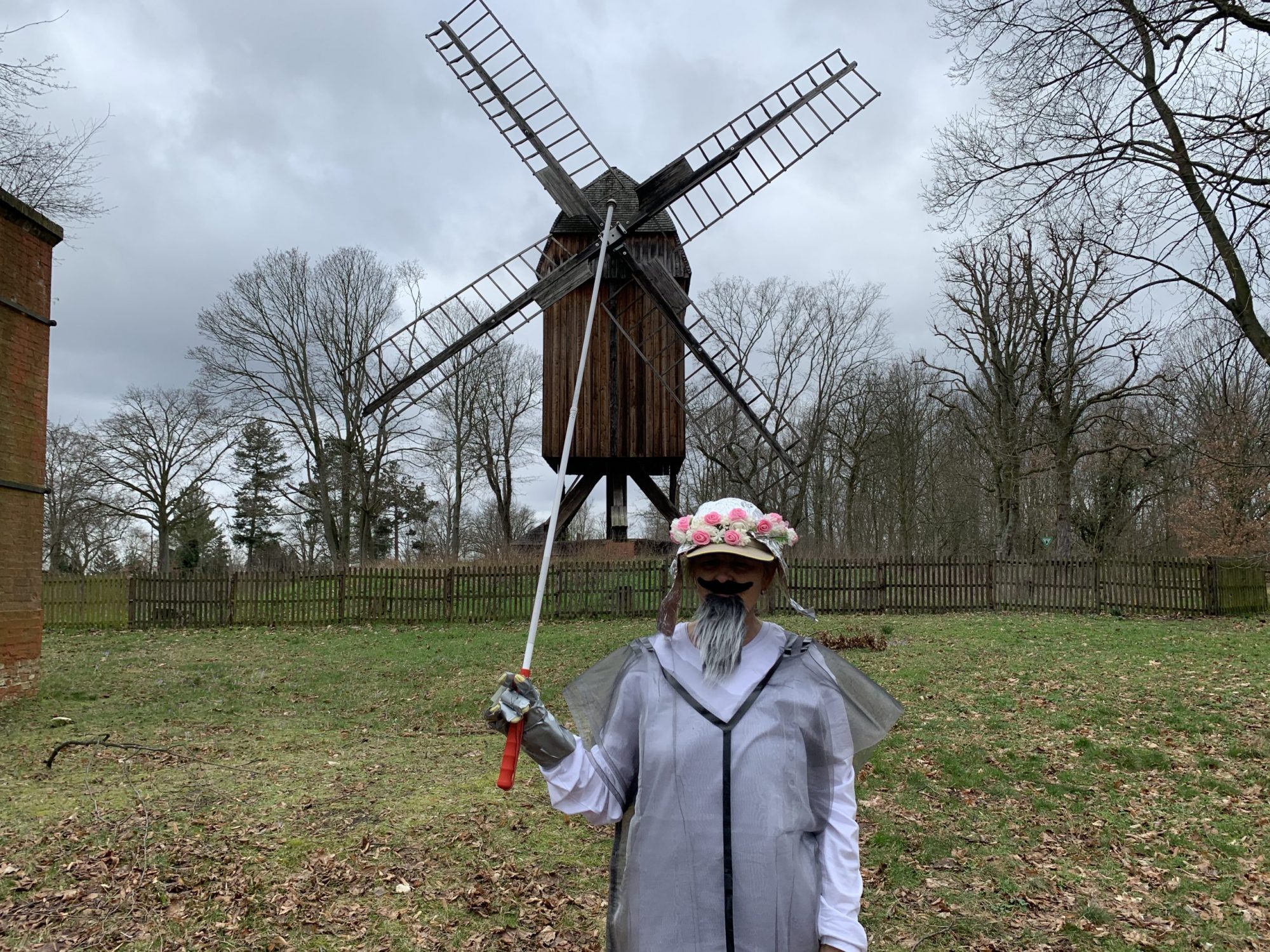 Die Blindgängerin als Don Quijote verkleidet auf einer Wiese. Sie hat einen grauen Bart und trägt einen Hut aus Alufolie mit einem Blütenkranz. Ihren weißen Langstock hält sie empor. Im Hintergrund eine Windmühle auf einem Grashügel.