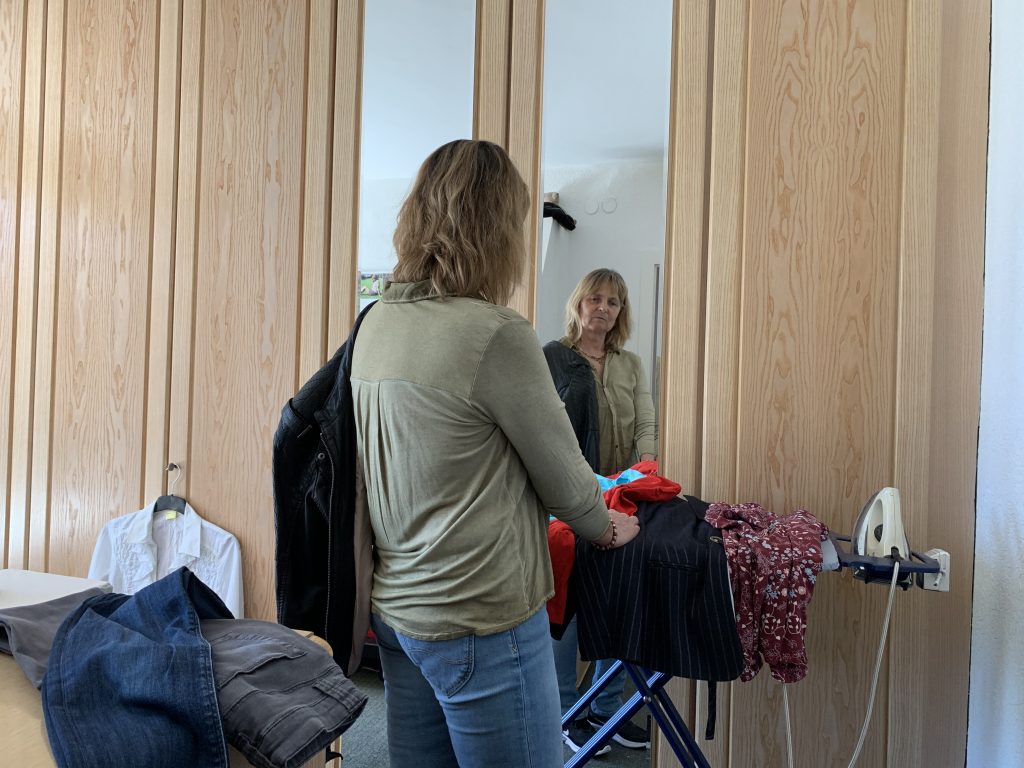 Die Blindgängerin vor einem Bügelbrett, das vor den Spiegeltüren eines Kleiderschrankes steht. Sie trägt eine olivfarbene Bluse zu hellen Jeans. Auf dem Bügelbrett diverse Kleidungsstücke in vielen Farben.