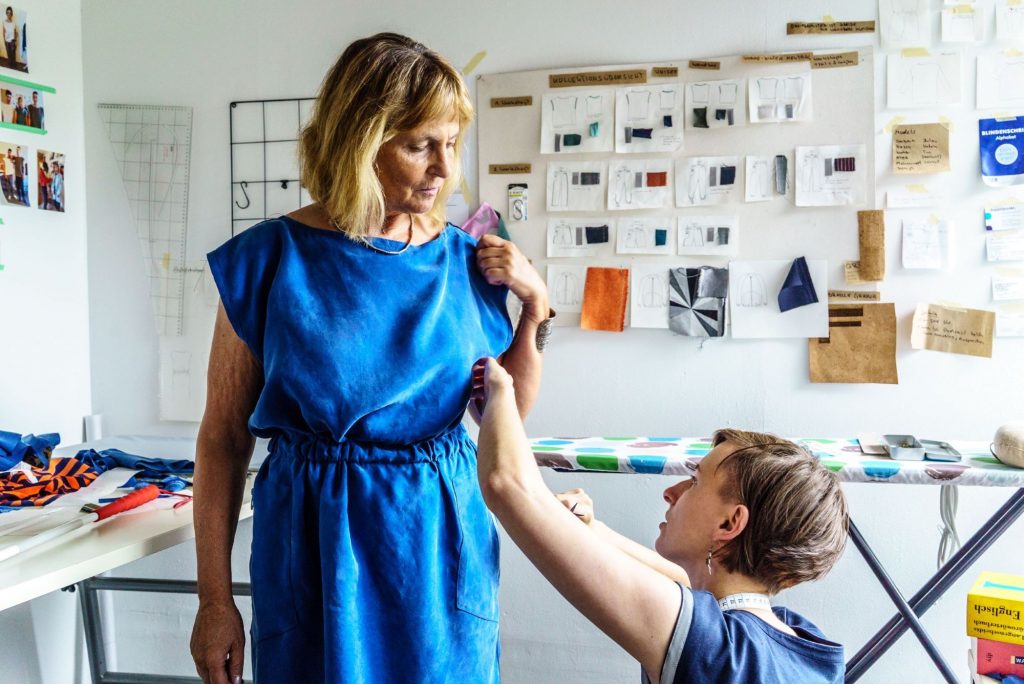 Die Blindgängerin steht in einem leuchtend blauen ärmellosen Kleid vor einem Bügelbrett. Eine Frau, Anna Flemmer, korrigiert den Sitz des Kleides.