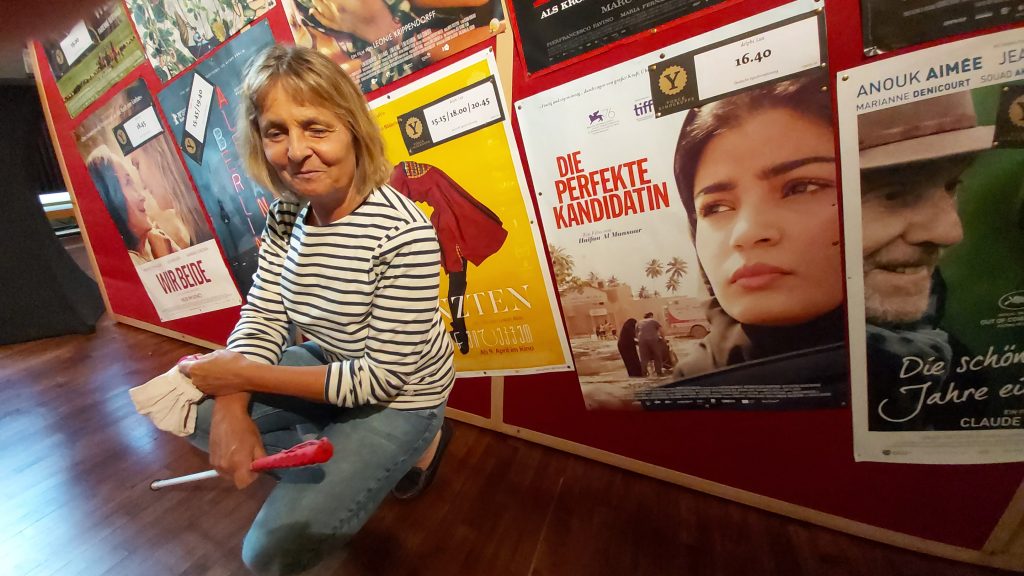 Die Blindgängerin in Jeans und geringeltem Shirt in der Hocke vor einer Wand mit Filmplakaten. In den Händen hält sie den weißen Stock und eine Atemschutzmaske. Rechts neben ihr das Plakat zum Film "Die perfekte Kandidatin".