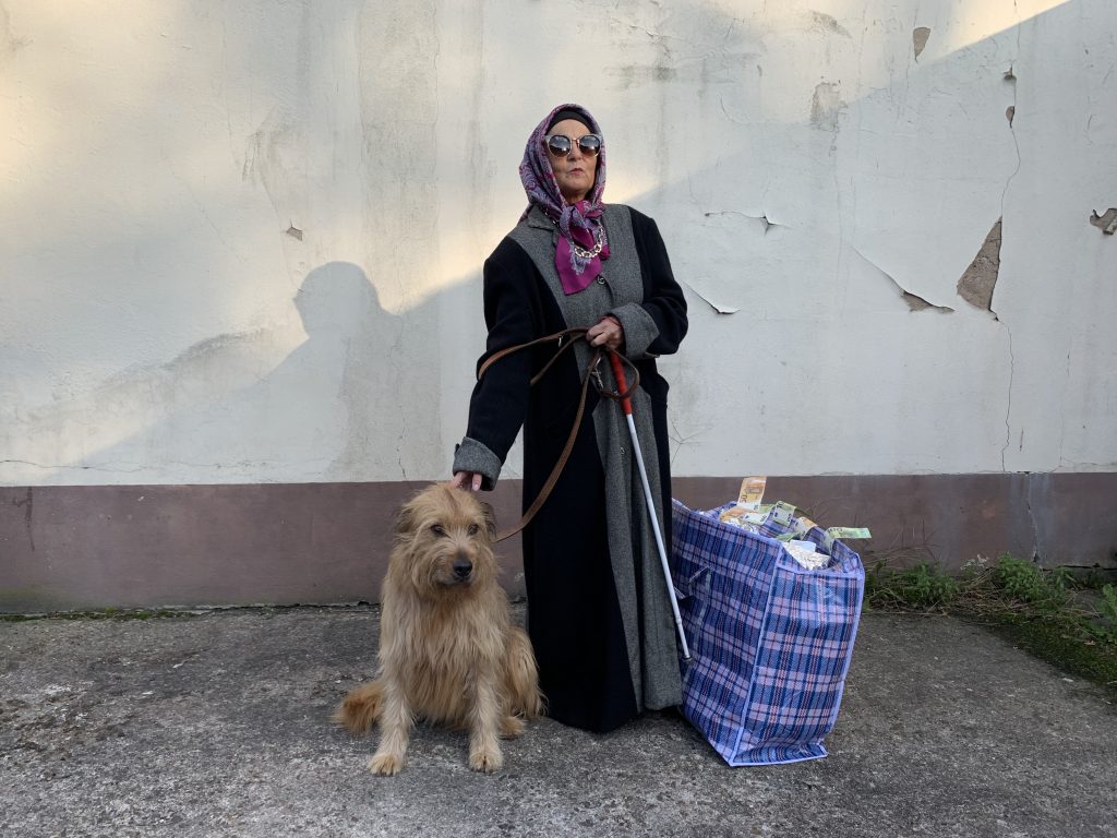 Die Blindgängerin vor einer weißen Fassade, an der der Putz bröckelt. Sie trägt einen bunten Hidschab, einen langen schwarzen Mantel, und hält ihren weißen Langstock. Neben ihr sitzt ein Hund, dem sie den Kopf tätschelt. Auf der anderen Seite steht eine große Plastiktasche am Boden, gefüllt mit in Alufolie gewickelten Päckchen. Geldscheine quillen aus der Tasche.