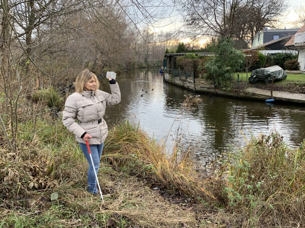 Die Blindgängerin steht am Ufer eines schmalen Kanals, auf dem Enten schwimmen. Sie trägt eine helle dicke Winterjacke. In der linken Hand hält sie einen Stein. Sie hebt den Arm, bereit zum Wurf.