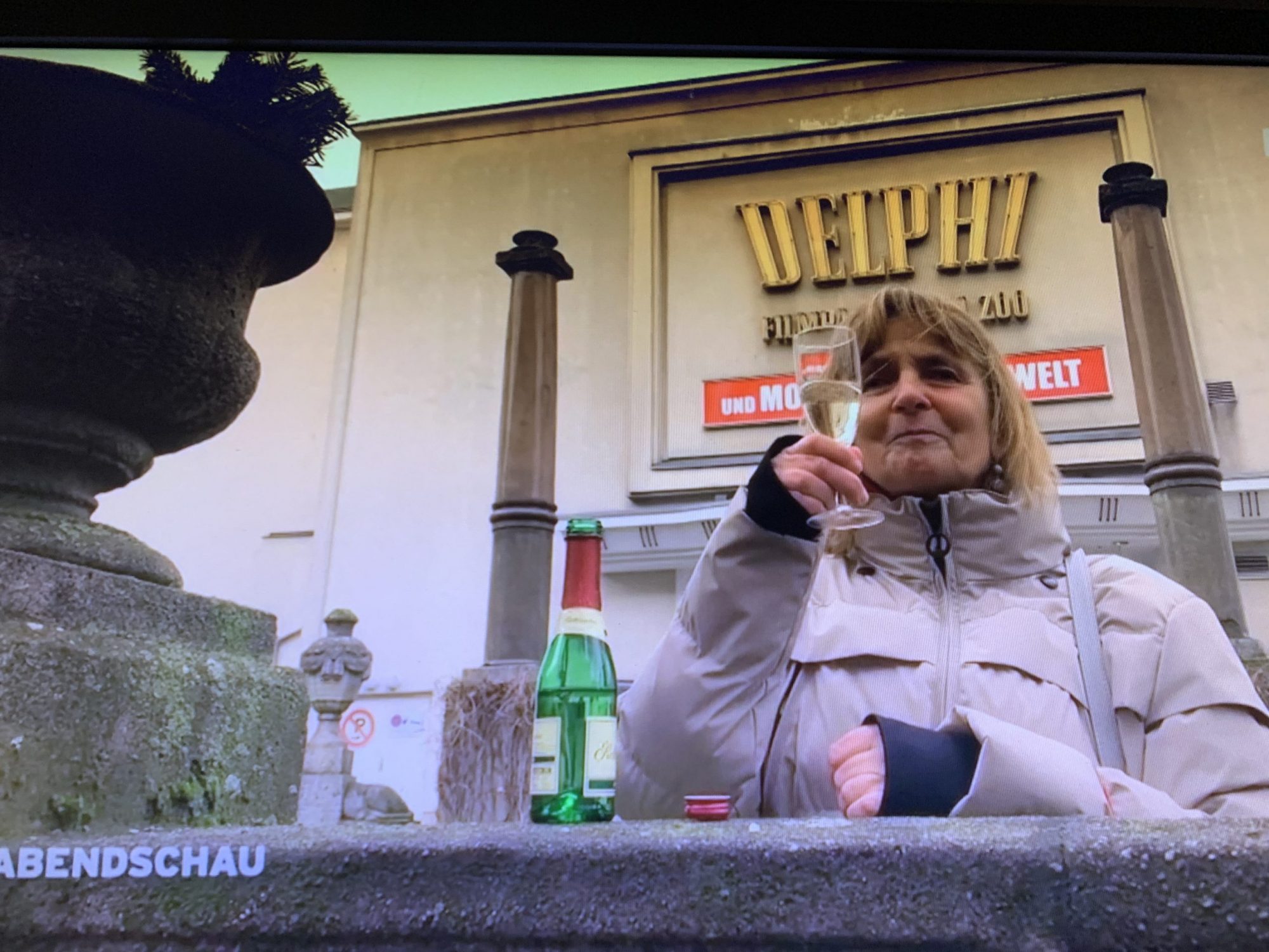 Vor dem Delphi-Filmpalast in Berlin steht die Blindgängerin in einer hellen dicken Winterjacke. Auf einer steinernen Balustrade vor ihr steht eine geöffnete Piccolo-Flasche. Die Blindgängerin hebt ein Glas Sekt hoch und prostet fröhlich der Kamera zu.