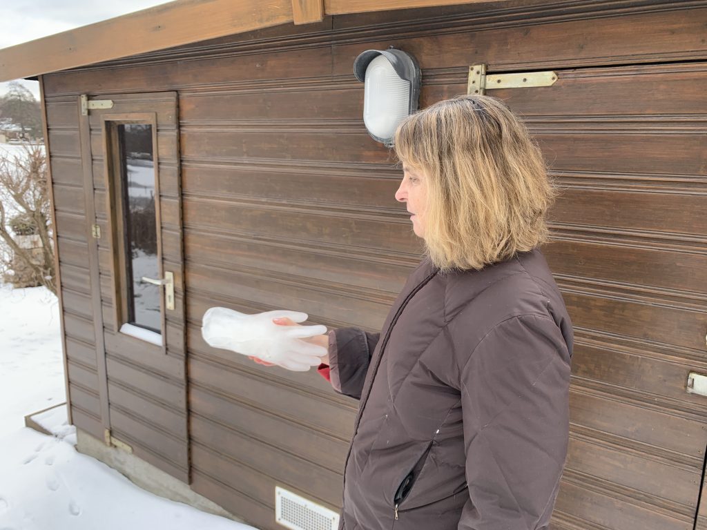 Vor einer braunen Holzhütte steht die Blindgängerin im Schnee. In ihrer ausgestreckten rechten Hand hält sie eine Hand aus Eis. Die Hände liegen wie bei einer Begrüßung ineinander.