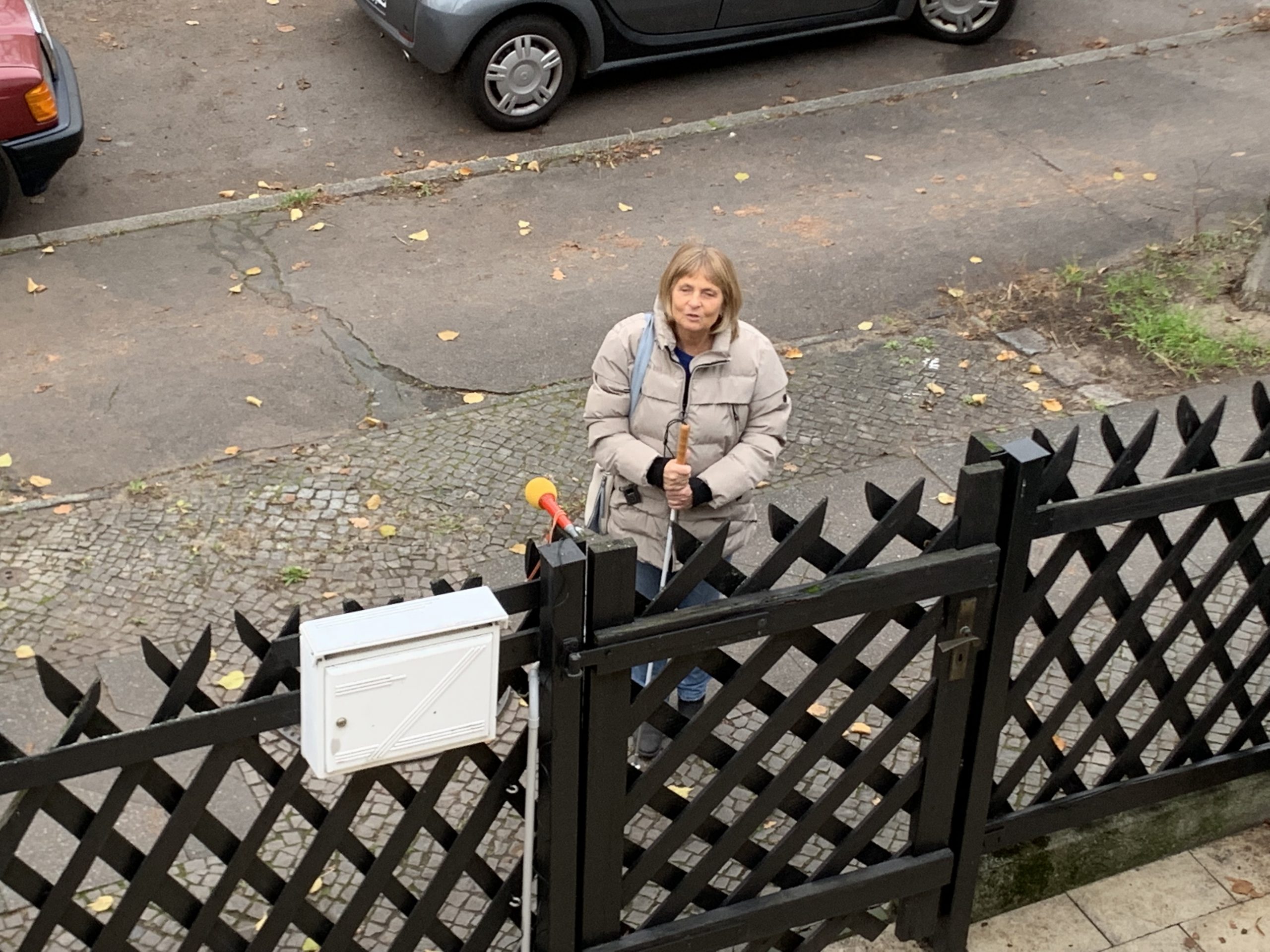 Die Blindgängerin von oben aufgenommen. In einer hellen Winterjacke steht sie auf dem Gehweg vor einem Gartenzaun. Am Zaunpfosten ist ein Mikrofon befestigt.