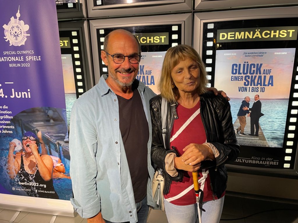 Der Hauptdarsteller Bernard Campan und die Blindgängerin stehen vor einer Wand mit Filmplakaten. Er legt seine Hand auf ihre Schulter. Neben ihnen das Plakat zum Film "Glück auf einer Skala von 1 bis 10".