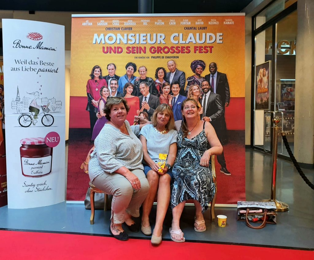 Die Blindgängerin mit ihren Freundinnen Astrid und Pascale auf einer Bank neben einem roten Teppich. Direkt hinter ihnen das Filmplakat "Monsieur Claude und sein großes Fest". Auf dem Plakat die große Filmfamilie. Links daneben eine Werbetafel mit dem Bild eines Marmeladenglases.