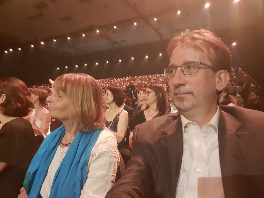 Die Blindgängerin mit ihrem Begleiter Andreas. Sie sitzen im Publikum zwischen vollbesetzten Stuhlreihen im Saal des Palais am Funkturm in Berlin.