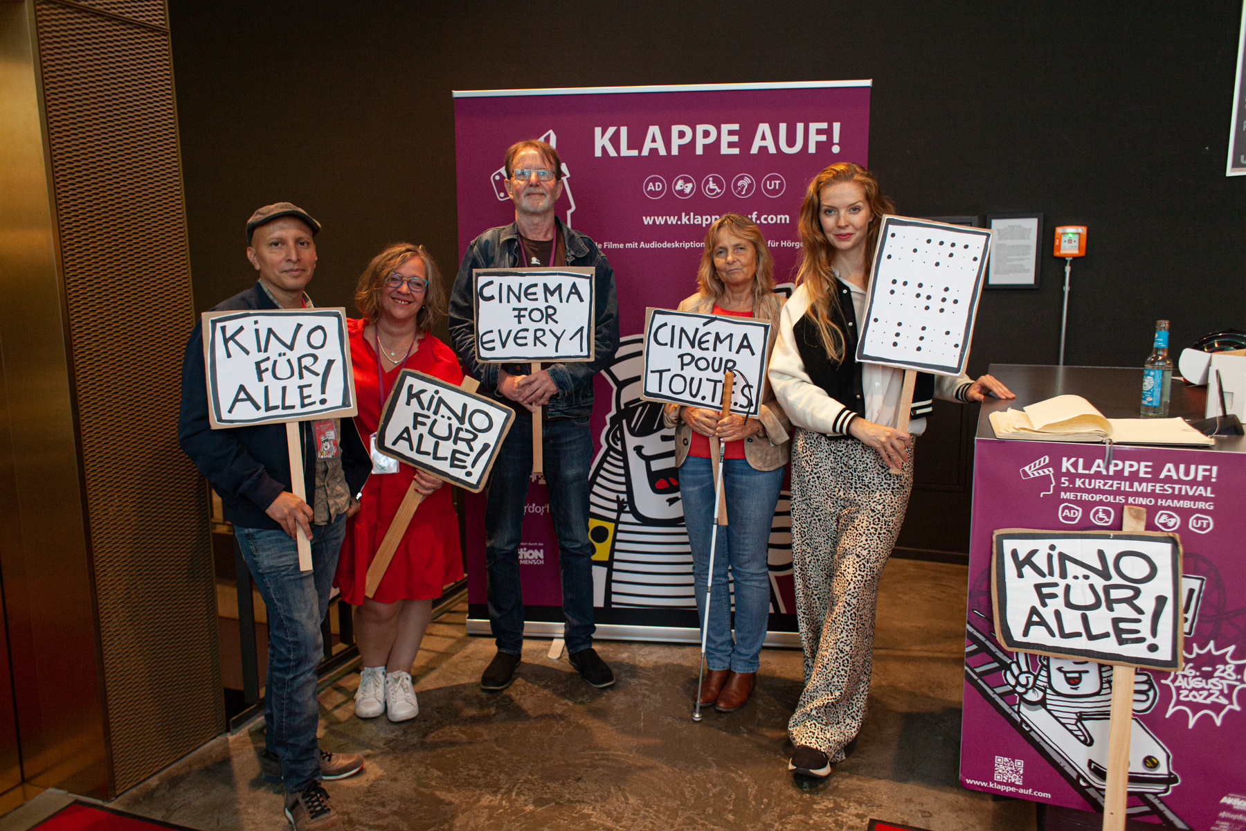 Im Foyer des Metropolis Kinos Hamburg hängen Plakate des KLAPPE AUF! Kurzfilmfestivals. Davor stehen nebeneinander die fünf Mitglieder der Jury. Jedes Mitglied hält ein Schild Richtung Kamera. Auf den Schildern steht in verschiedenen Sprachen: Kino für alle!