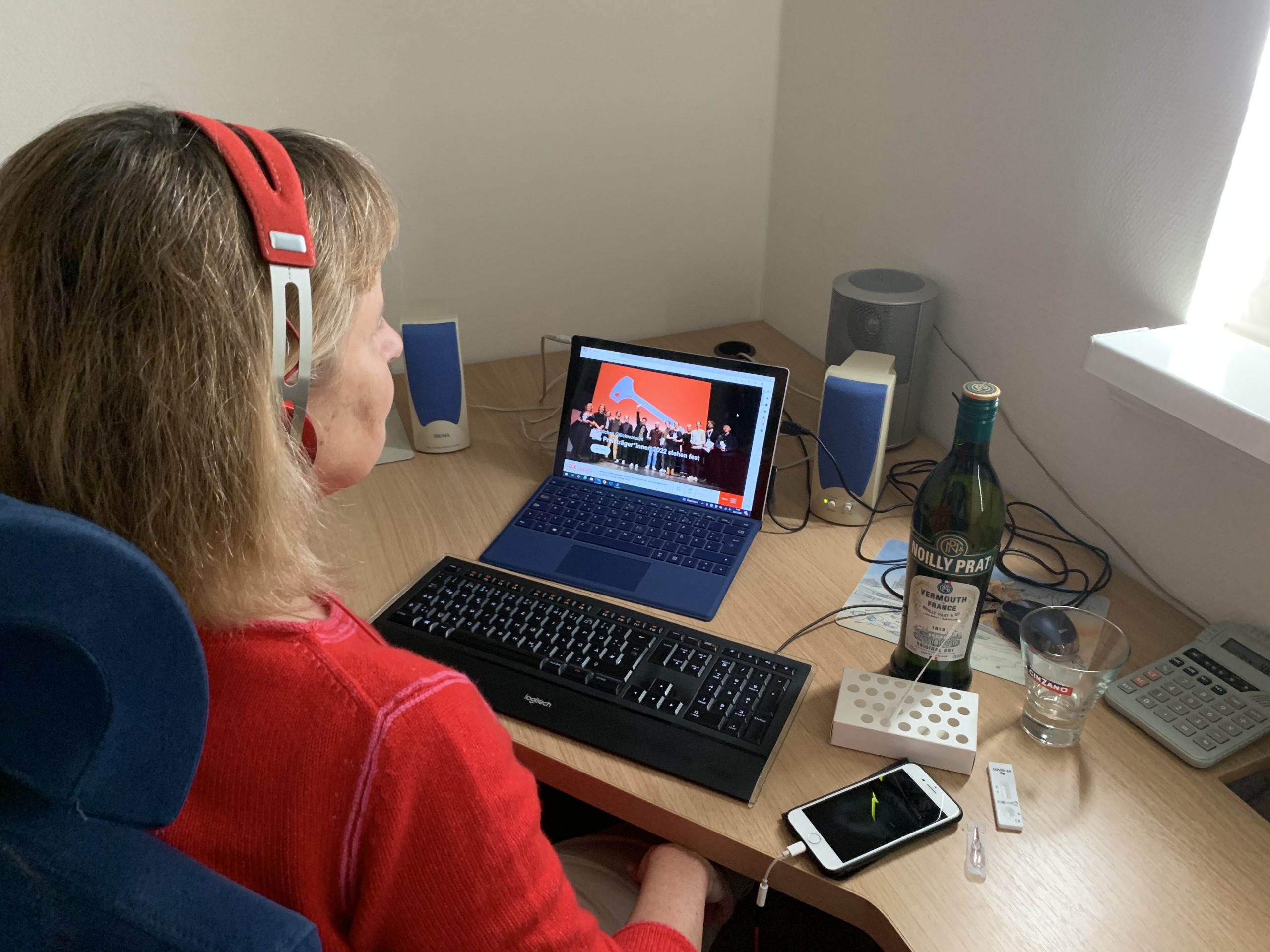 Im roten Pulli sitzt die Blindgängerin mit Kopfhörern an ihrem Schreibtisch, auf dem Bildschirm ihres Laptops die Startseite des DOK Leipzig. Links und rechts stehen Lautsprecherboxen. Rechts neben der Tastatur liegt ein Smartphone, daneben steht eine Flasche Wermut mit einem Glas. Ein ausgepacktes Corona-Testset liegt auf dem Tisch.