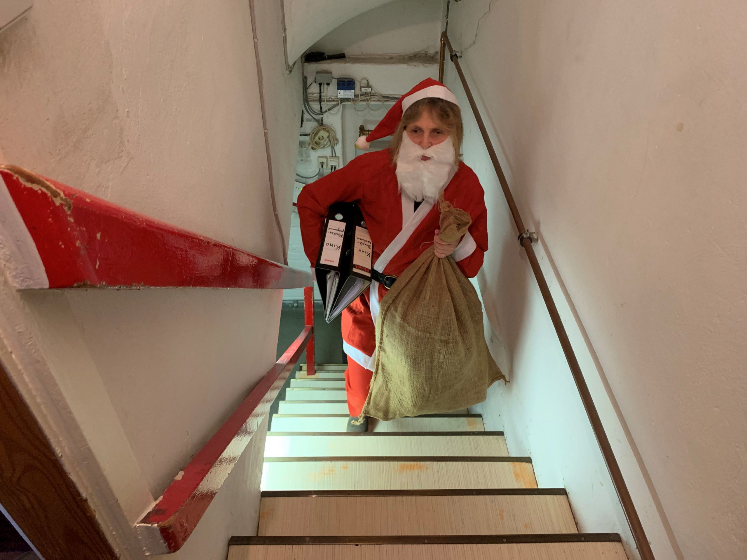 Die Blindgängerin im roten Weihnachtsmannkostüm und mit weißem Bart steigt eine schmale Kellertreppe empor. In der linken Hand hält sie einen vollen braunen Sack, unter dem rechten Arm klemmen zwei Aktenordner.