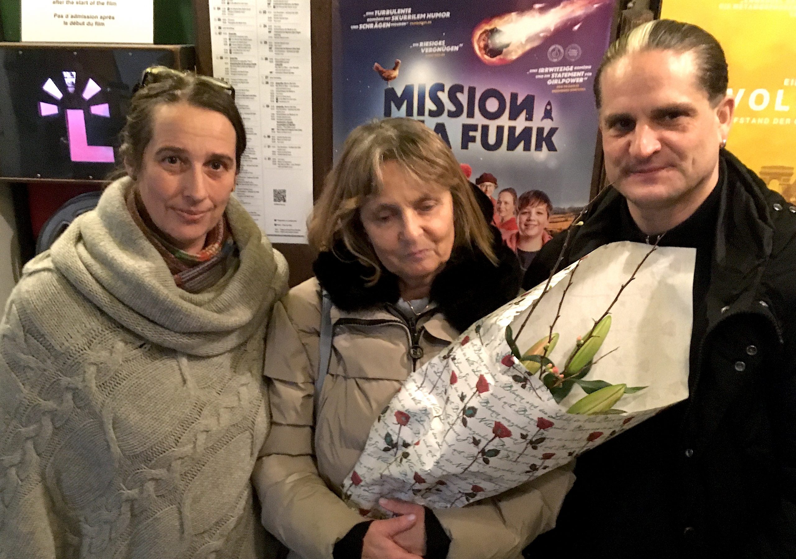 Die Autorin von Audiodeskriptionen Jutta Polić, die Blindgängerin und Andres Schüpdach von der Greta App stehen vor einer Wand im Lichtblick-Kino. Hinter ihnen das Filmplakat von „Mission Ulja Funk“. Die Blindgängerin hält einen großen Blumenstrauß im Arm.