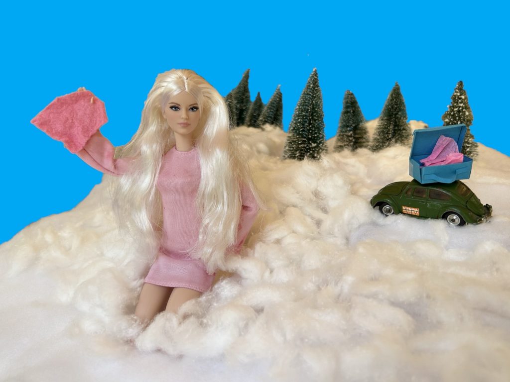 Eine Barbie-Puppe mit langen blonden Haaren sitzt im tiefen Schnee, der aus Watte ist. Sie trägt ein pinkfarbenes Kleid, schaut in die Kamera und winkt mit einem pinkfarbenen Tuch. Auf dem verschneiten Berg hinter ihr ein grüner VW-Käfer. Auf seinem Dach steht ein geöffneter blauer Koffer. Am Gipfel des Watte-Schneeberges stehen grüne Tannen mit Schnee in den Zweigen. Dahinter ein klarer blauer Himmel.