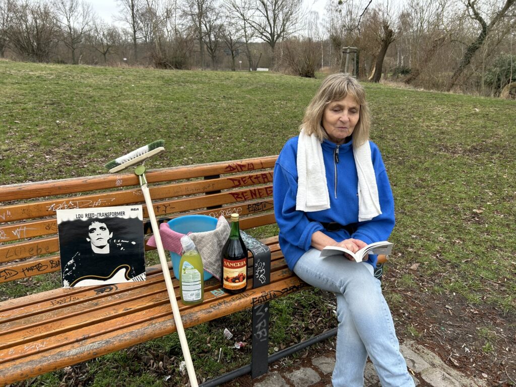 Die Blindgängerin sitzt auf einer Parkbank auf einer mit Bäumen und Sträuchern umgebenen Wiese und schaut in ein Taschenbuch. Sie trägt einen blauen Pullover mit einem schwarzen Gürtel und blaue Jeans. Ein weißes Handtuch ist um die Schultern gelegt. Neben ihr auf der Bank ein Eimer mit Putzlappen, WC-Reiniger, ein Schrubber und eine große Flasche Sangria. An der Rückenlehne der Bank steht die LP "Transformer" von Lou Reed.